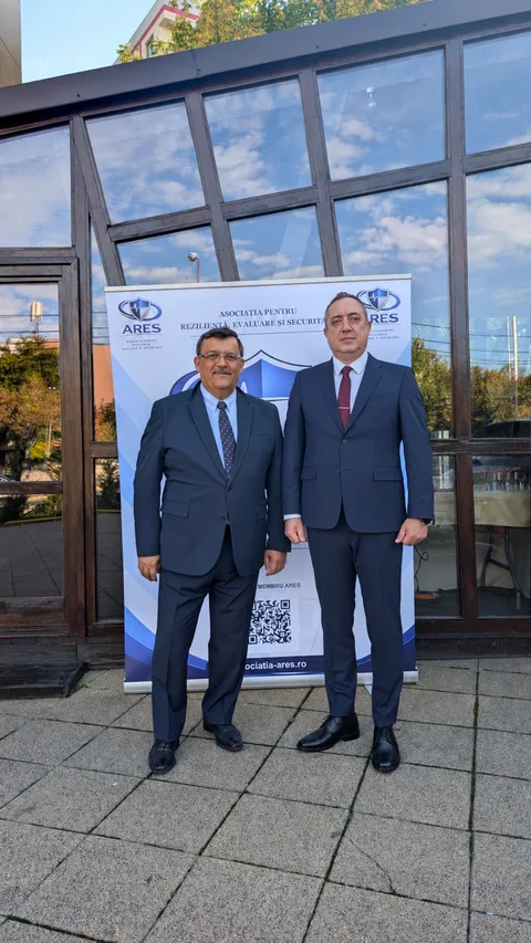 Doi participanți în costume, fotografiați în exterior lângă bannerul ARES