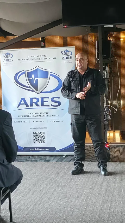 Participant în jachetă închisă la culoare, adresându-se sălii la Forumul ARES